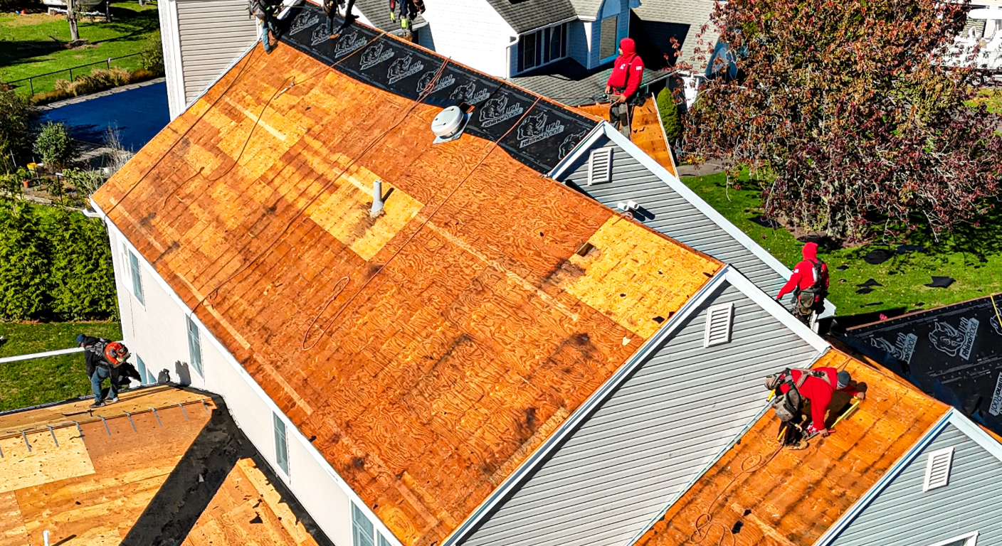 A professional roofing crew works on a large, grey suburban home under a bright blue sky.