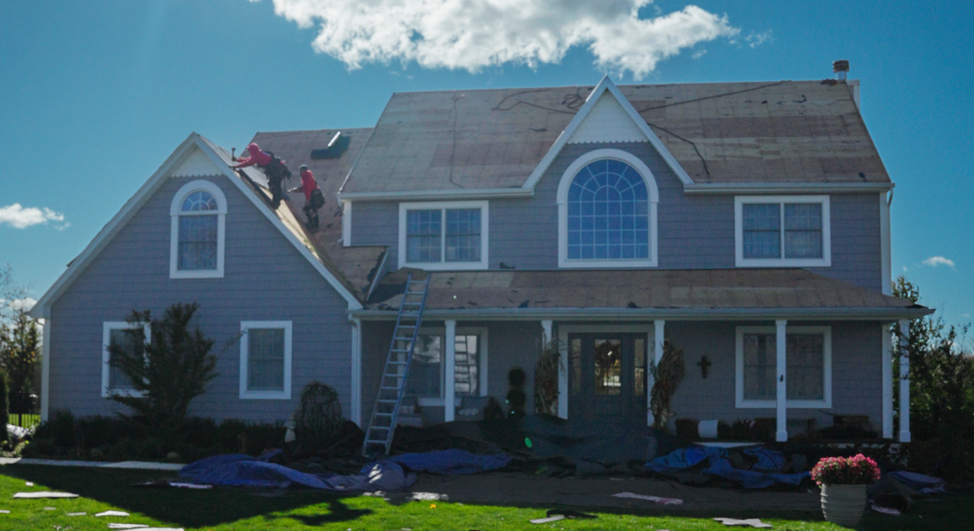 A roofer in a red hoodie and safety harness climbs a ladder to install shingles.