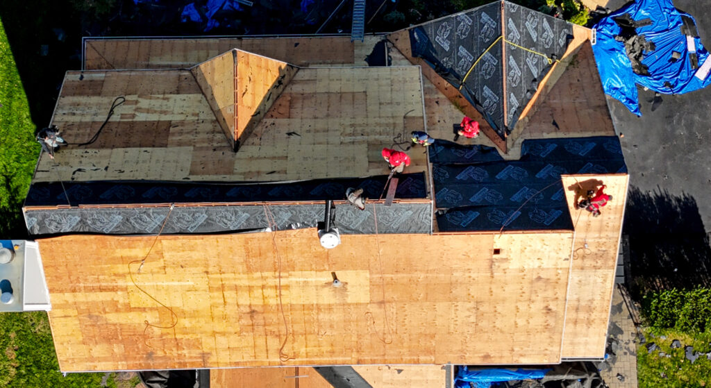 A roofer in a red hoodie and safety harness climbs a ladder to install new shingles.