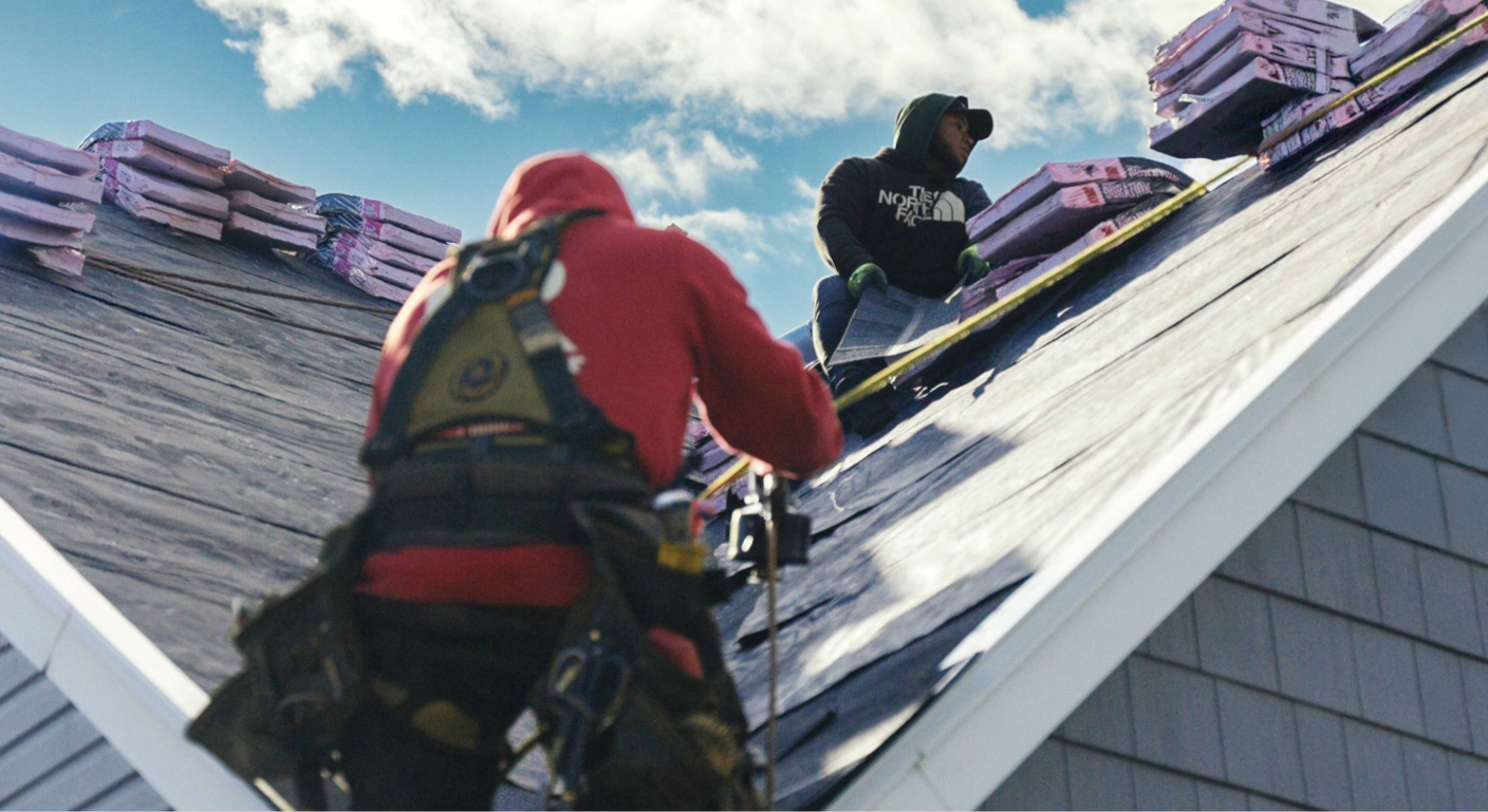 Two roofers secured by safety ropes carefully strip old materials from a steep residential roof valley.