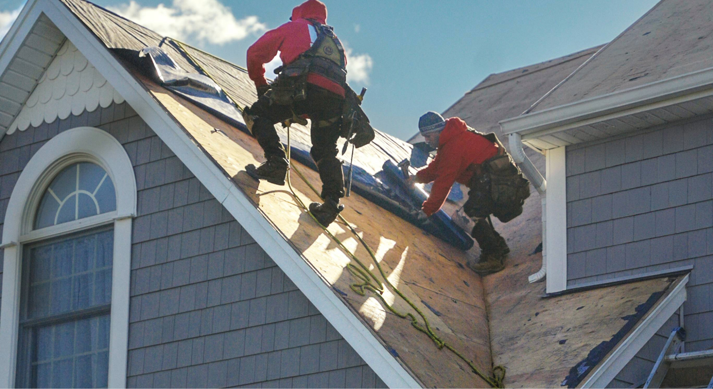 A professional roofing crew works on a large, grey suburban home under a bright blue sky.