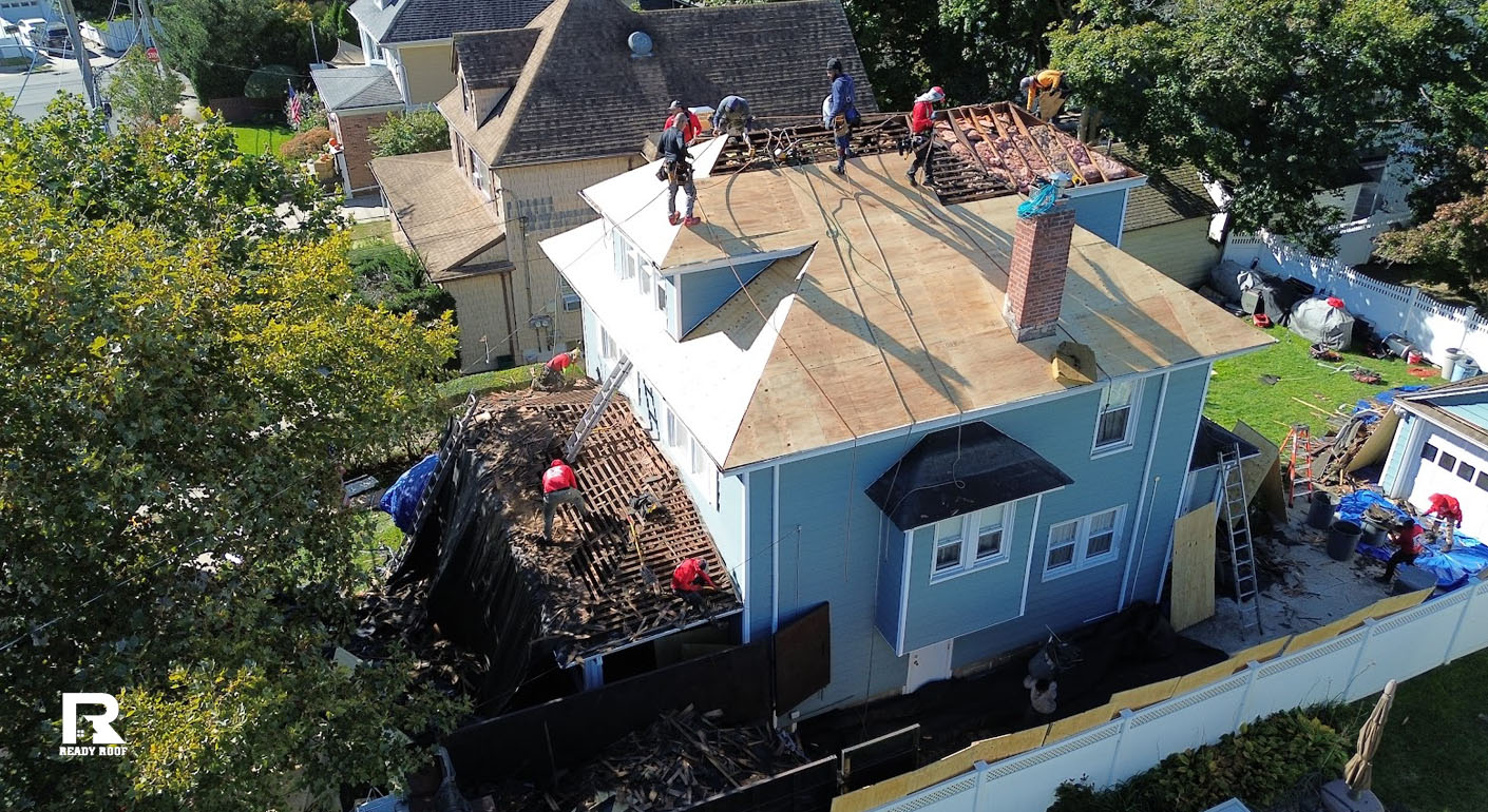 Aerial view showing workers removing an old roof on a blue house, considering metal roofing vs asphalt shingles for replacement.
